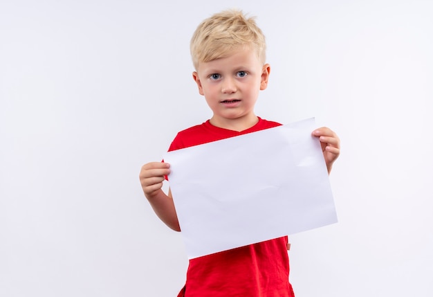 Um menino loiro fofo com uma camiseta vermelha segurando uma folha de papel em branco enquanto olha para uma parede branca