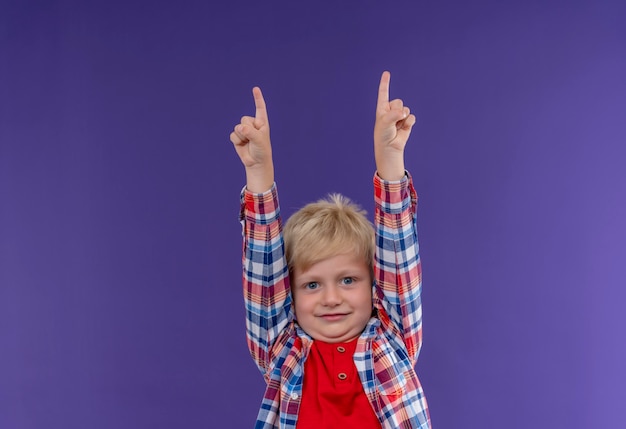 Um menino sorridente e fofo com cabelo loiro vestindo uma camisa xadrez apontando para cima com os dedos indicadores olhando para uma parede roxa