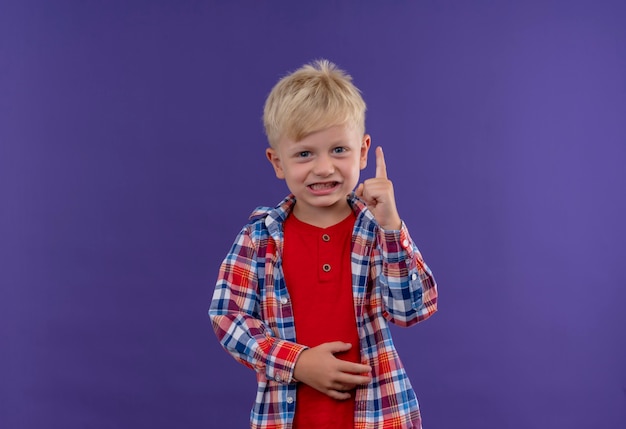 Foto grátis um menino sorridente e fofo com cabelo loiro vestindo uma camisa xadrez levantando o dedo indicador enquanto olha para uma parede roxa