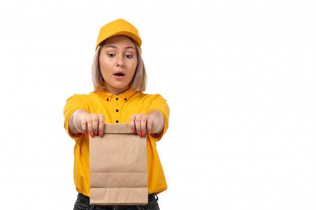 Um mensageiro feminino vista frontal na camisa amarela, boné amarelo e calça jeans preta, segurando o pacote de alimentos em branco