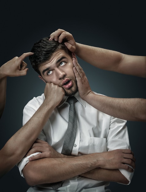 Um modelo masculino rodeado por mãos como seus próprios pensamentos na parede escura. Um jovem duvida, não consegue escolher a decisão certa. Conceito de problemas mentais, problemas no trabalho, indecisão.