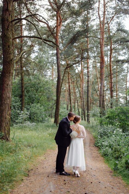 Um noivo barbudo e elegante de terno e uma linda noiva loira em um vestido branco com um buquê nas mãos estão de pé e se abraçando na natureza na floresta de pinheiros.