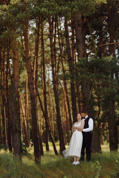 Foto grátis um noivo barbudo e elegante de terno e uma linda noiva loira em um vestido branco com um buquê nas mãos estão de pé e se abraçando na natureza na floresta de pinheiros.
