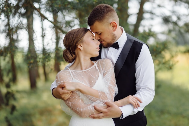 Um noivo barbudo e elegante de terno e uma linda noiva loira em um vestido branco com um buquê nas mãos estão de pé e se abraçando na natureza na floresta de pinheiros.