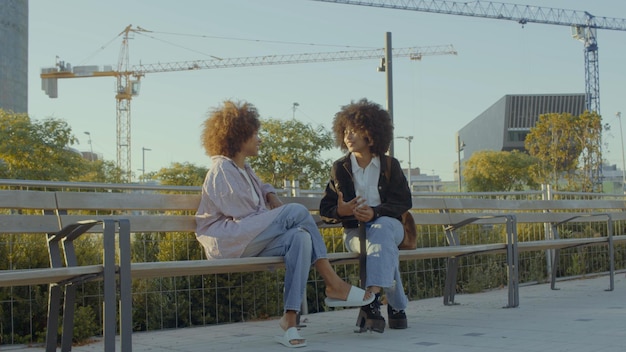 Um par de mulheres negras de raça mista no parque sentadas e conversando com edifícios da cidade no fundo