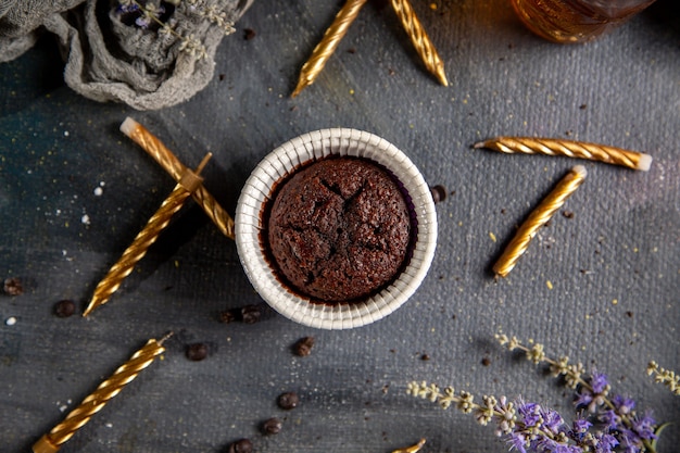 Um pequeno bolo de chocolate com velas, flores roxas e chá na mesa cinza.