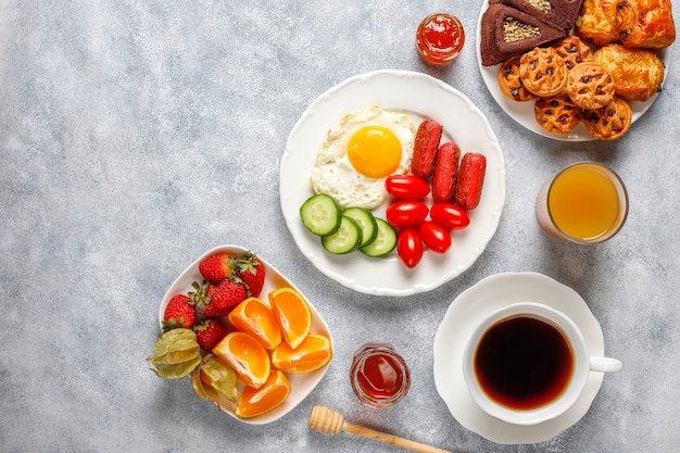 Um prato de café da manhã contendo linguiças de coquetel, ovos fritos, tomate cereja, doces, frutas e um copo de suco de pêssego.