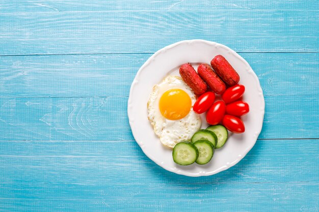 Um prato de café da manhã contendo linguiças de coquetel, ovos fritos, tomate cereja, doces, frutas e um copo de suco de pêssego.