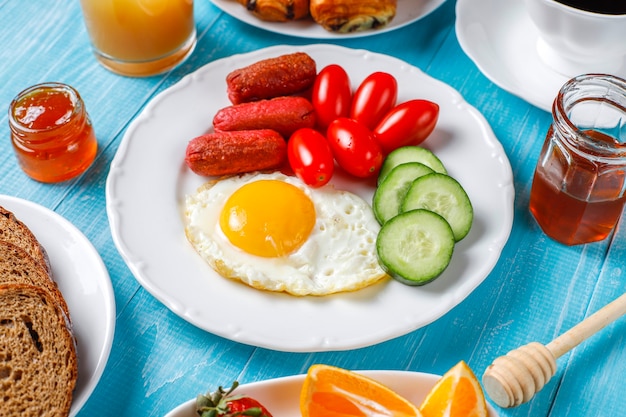 Um prato de café da manhã contendo linguiças, ovos fritos, tomate cereja, doces, frutas e um copo de suco de pêssego.