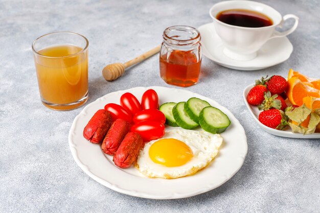 Um prato de café da manhã contendo linguiças, ovos fritos, tomate cereja, doces, frutas e um copo de suco de pêssego.