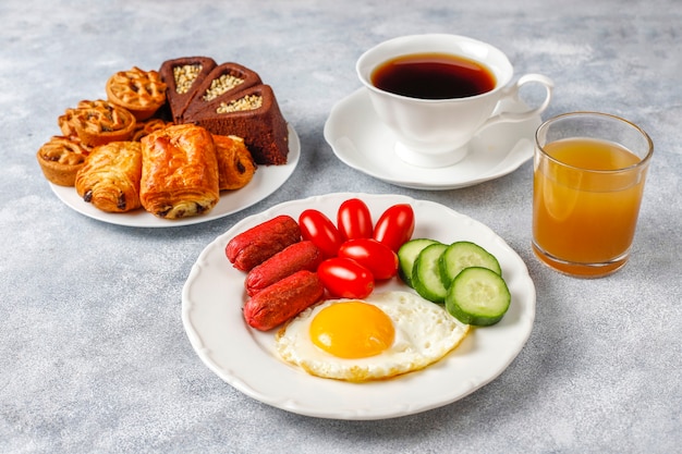 Um prato de café da manhã contendo linguiças, ovos fritos, tomate cereja, doces, frutas e um copo de suco de pêssego.