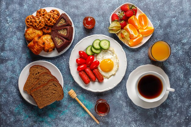 Um prato de café da manhã contendo linguiças, ovos fritos, tomate cereja, doces, frutas e um copo de suco de pêssego.