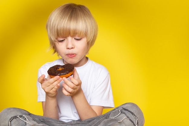 Foto grátis um rapaz bonito loiro vista frontal em t-shirt branca comendo rosquinhas na mesa amarela