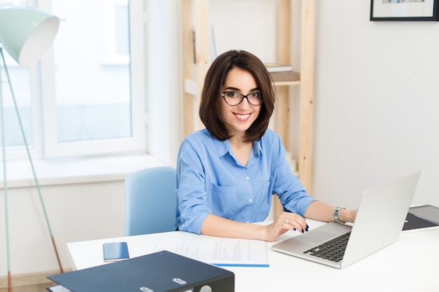 Um retrato de uma menina bonita sentada à mesa no escritório. Ela está trabalhando com o laptop e sorrindo para a câmera.