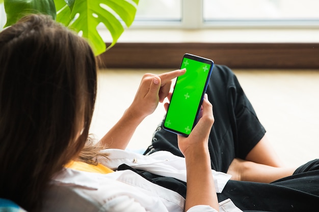 Um smartphone vertical com uma tela chromakey na mão de uma garota que está sentada perto da janela
