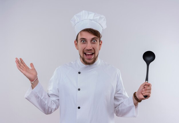 Um surpreendente jovem chef barbudo de uniforme branco segurando uma concha preta enquanto olha para uma parede branca