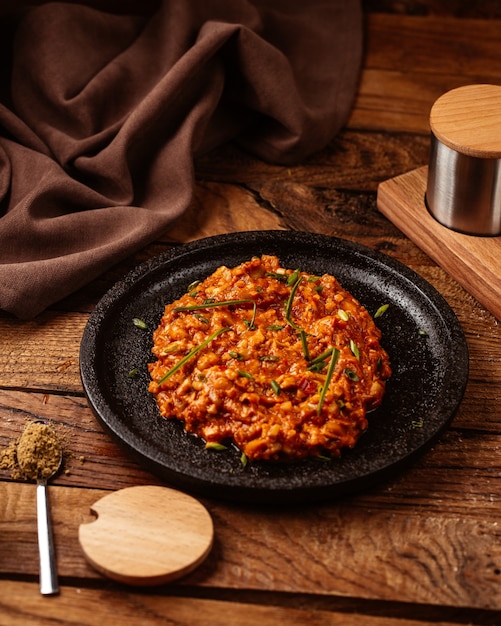 Um tomate de frente com carne picada frita e quente dentro de uma placa preta na mesa de madeira.