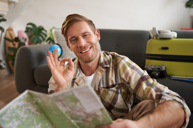Um turista bonito sorridente com um mapa de viagem sentado no chão perto de uma mala mostra o sinal ok ok