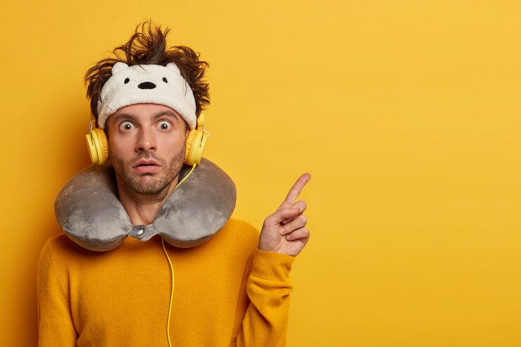 Foto grátis um turista chocado aponta para longe, mostra algo em um espaço em branco com estupefação, usa uma venda nos olhos e um travesseiro inflado em volta do pescoço