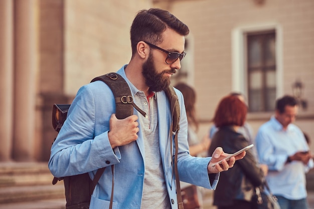Um turista com barba e cabelo cheios, vestindo roupas casuais e óculos de sol, segura uma mochila e mandando mensagens em um smartphone, em uma rua antiga, durante a excursão na Europa.