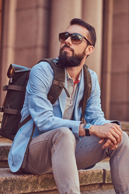 Foto grátis um turista com barba e corte de cabelo cheio, vestindo roupas casuais com mochila e óculos de sol, sentado em um degrau em uma rua antiga.