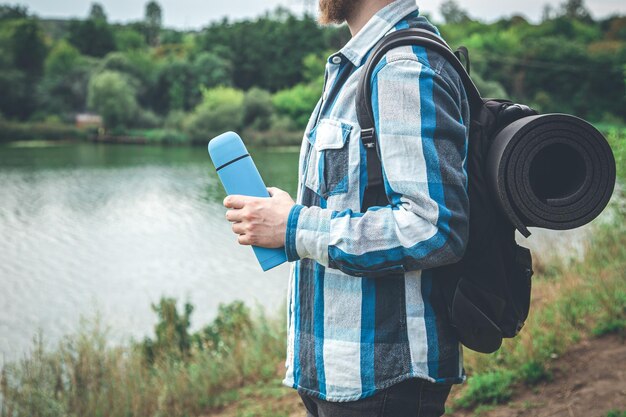 Um viajante de homem com uma mochila e um karemat segura uma garrafa térmica na mão