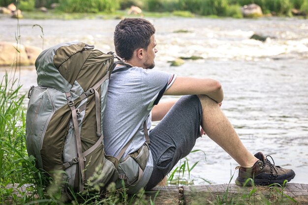 Um viajante do sexo masculino com uma mochila grande para caminhadas descansa perto do rio.