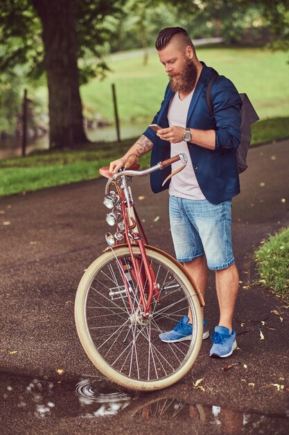 Um viajante vestido com roupas casuais com uma mochila, usando um smartphone, relaxando em um parque da cidade depois de andar de bicicleta retrô.