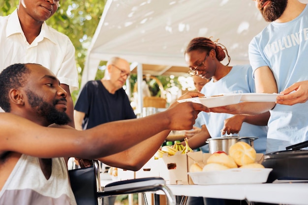 Um voluntário ajuda e fornece comida grátis a um homem afro-americano pobre e necessitado em cadeira de rodas voluntários em um banco de alimentos ao ar livre fornecem assistência à fome aos aleijados e desfavorecidos