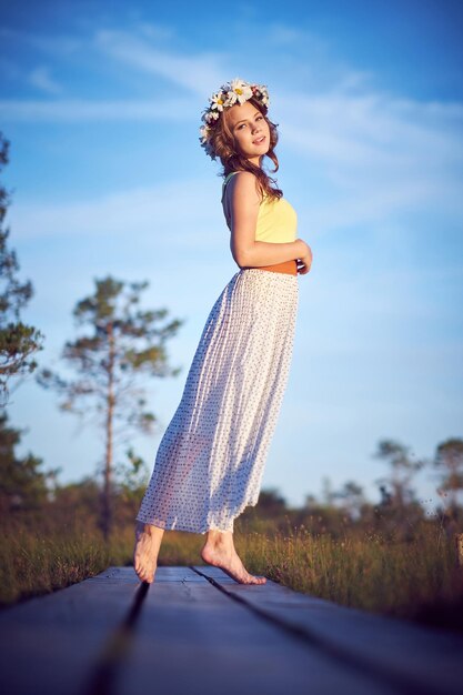 Uma adolescente atraente vestida com uma saia longa e guirlanda na cabeça posando no campo ao nascer do sol.