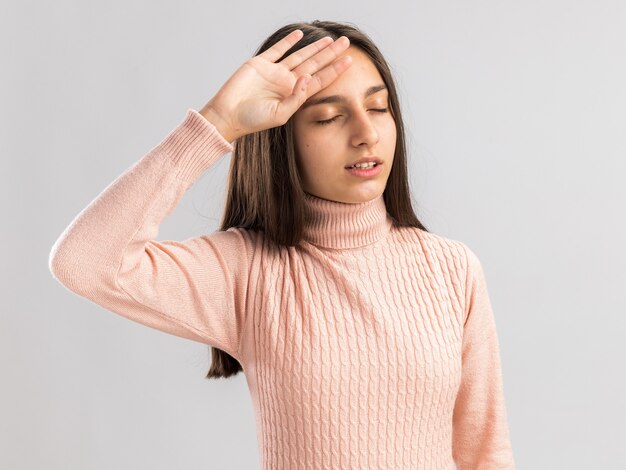 Uma adolescente bonita cansada mantendo a mão na cabeça com os olhos fechados, isolado na parede branca