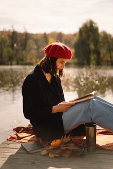 Uma adolescente com uma boina vermelha lendo um livro no pontão de madeira na temporada de outono