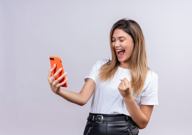 Uma adorável jovem muito feliz em uma camiseta branca levantando o punho cerrado enquanto segura o telefone