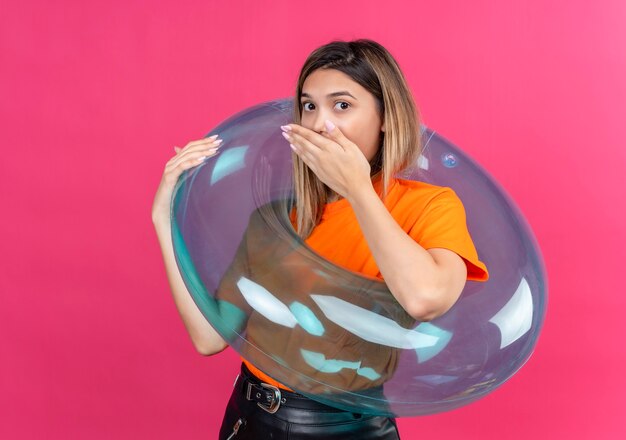 Uma adorável jovem surpresa em uma camiseta laranja, mantendo a mão na boca enquanto está de pé em um anel inflável em uma parede rosa