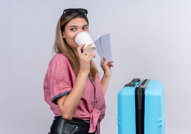 Uma adorável jovem vestindo camisa vermelha e óculos escuros, bebendo um café enquanto segura passagens aéreas em uma parede branca