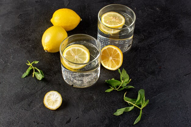 Uma água de vista frontal com bebida fresca de limão com limões fatiados, juntamente com limões inteiros e folhas dentro de vidros transparentes no escuro