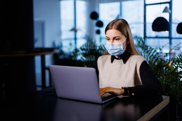 Uma bela empresária inteligente em uma grande empresa realiza uma reunião online com parceiros de negócios em uma máscara protetora durante o coronavírus, ela se senta em uma mesa em um local de trabalho com um laptop