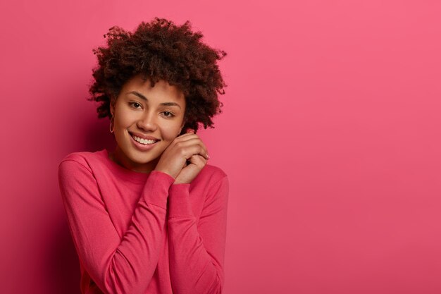 uma bela jovem afro-americana inclina a cabeça, mantém as mãos perto do rosto, tem um sorriso atraente, vestida com roupa casual, posa sobre uma parede rosa, espaço livre para seu anúncio
