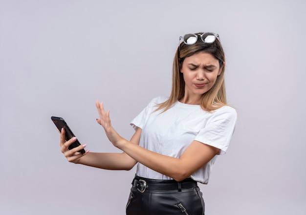 Uma bela jovem em uma camiseta branca usando óculos escuros na cabeça expressando negatividade enquanto segura o celular em uma parede branca