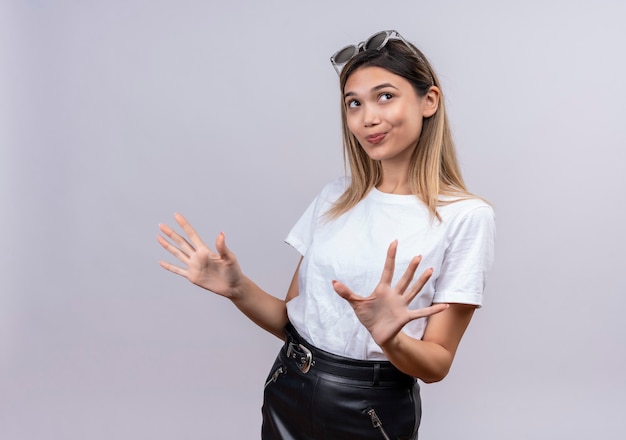 Foto grátis uma bela jovem sorridente com camiseta branca e óculos escuros na cabeça expressando positividade enquanto pensava em uma parede branca