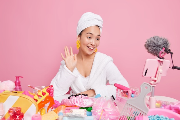 Uma blogueira asiática de beleza feminina sorridente filmes tutorial de maquiagem acenando olá na câmera do smartphone