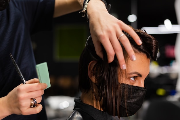 Uma cabeleireira profissional faz o corte de cabelo de um cliente. A menina está sentada com uma máscara no salão de beleza