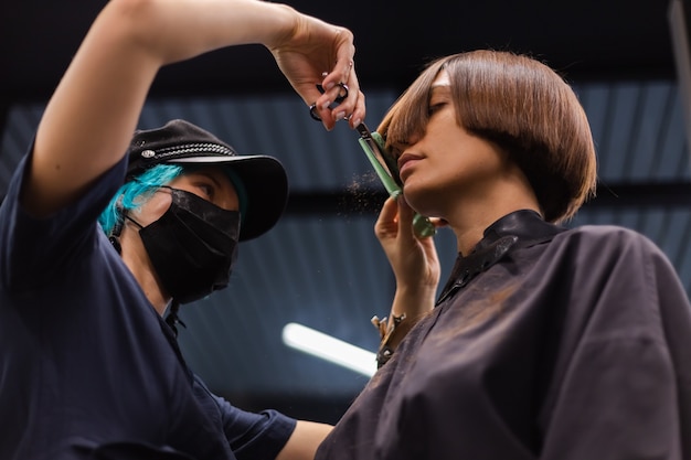 Foto grátis uma cabeleireira profissional faz o corte de cabelo de um cliente. a menina está sentada com uma máscara no salão de beleza