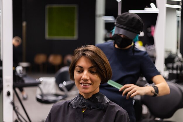 Uma cabeleireira profissional faz o corte de cabelo de um cliente. A menina está sentada com uma máscara no salão de beleza