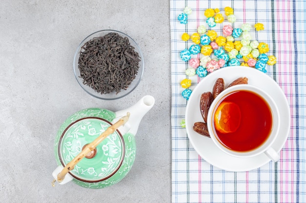 Uma composição de bule, uma pequena tigela de folhas de chá e uma xícara de chá em uma toalha com doces de pipoca espalhados no fundo de mármore. Foto de alta qualidade