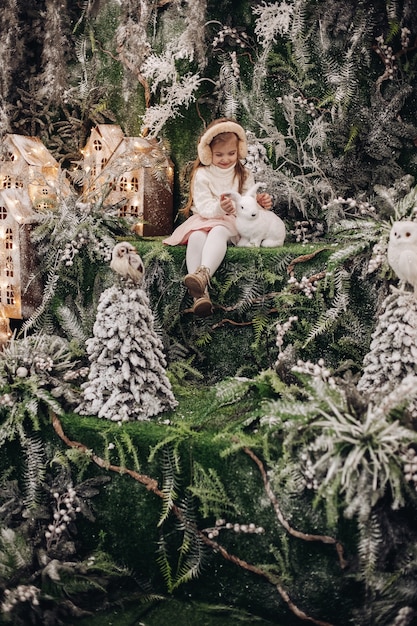 Uma criança bonita caucasiana com longos cabelos loiros sentada em uma atmosfera de natal com várias árvores decoradas ao seu redor e um coelhinho
