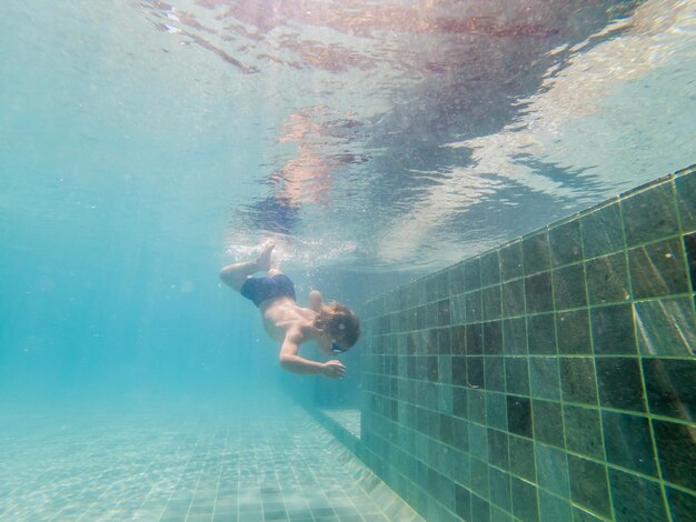 Uma criança menino está nadando debaixo d'água em uma piscina sorrindo e prendendo a respiração com óculos de natação