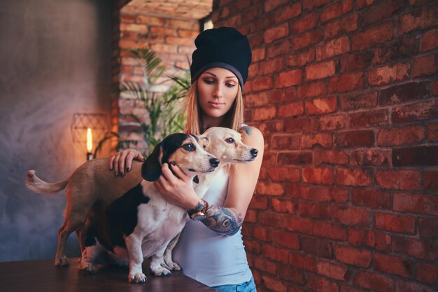 Uma elegante mulher loira tatuada em t-shirt e jeans abraça dois cachorros fofos.