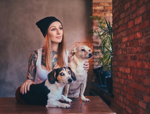 Uma elegante mulher loira tatuada em t-shirt e jeans abraça dois cachorros fofos.