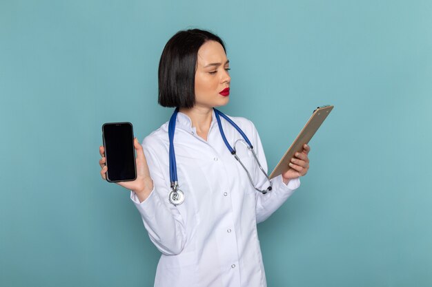 Uma enfermeira feminina jovem vista frontal no terno médico branco e estetoscópio azul segurando o bloco de notas e telefone